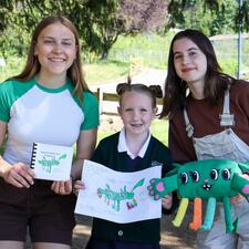 Two female secondary students stand with female elementary student holding their projects
